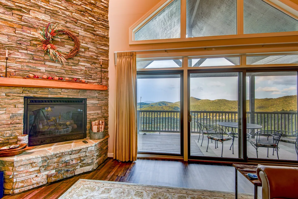 A warm, inviting living area featuring a stone fireplace, expansive windows with mountain views, polished hardwood floors, and tasteful decor. The space exudes a cozy charm, perfect for relaxing by the fire or enjoying the natural light that pours in.