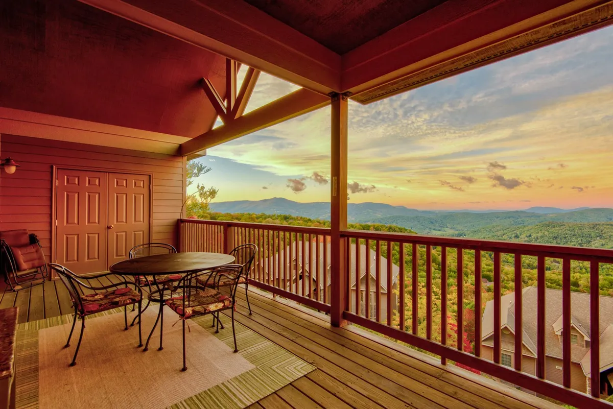 A serene outdoor patio set against the backdrop of the Blue Ridge Mountains, featuring comfortable patio furniture oriented towards a sweeping sunset sky. The view spreads out to reveal rolling peaks and vibrant sky colors, suggesting a tranquil space for outdoor dining or relaxing while soaking in the natural beauty.
