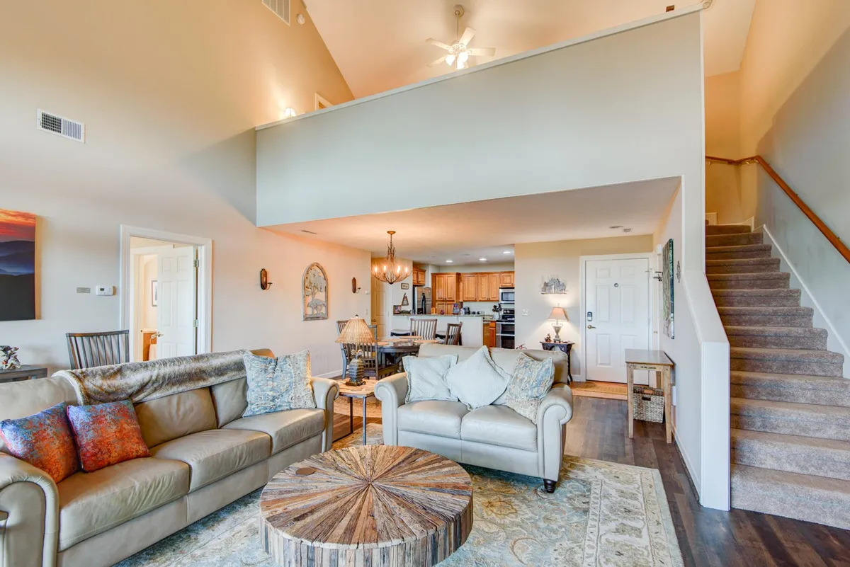 An inviting open-plan living and dining area featuring a comfortable sofa and coffee table, with a clear view of a modern kitchen highlighted by granite countertops and a stylish chandelier overhead. The staircase and spacious layout suggest easy movement between shared and private spaces, ideal for gatherings and family activities.