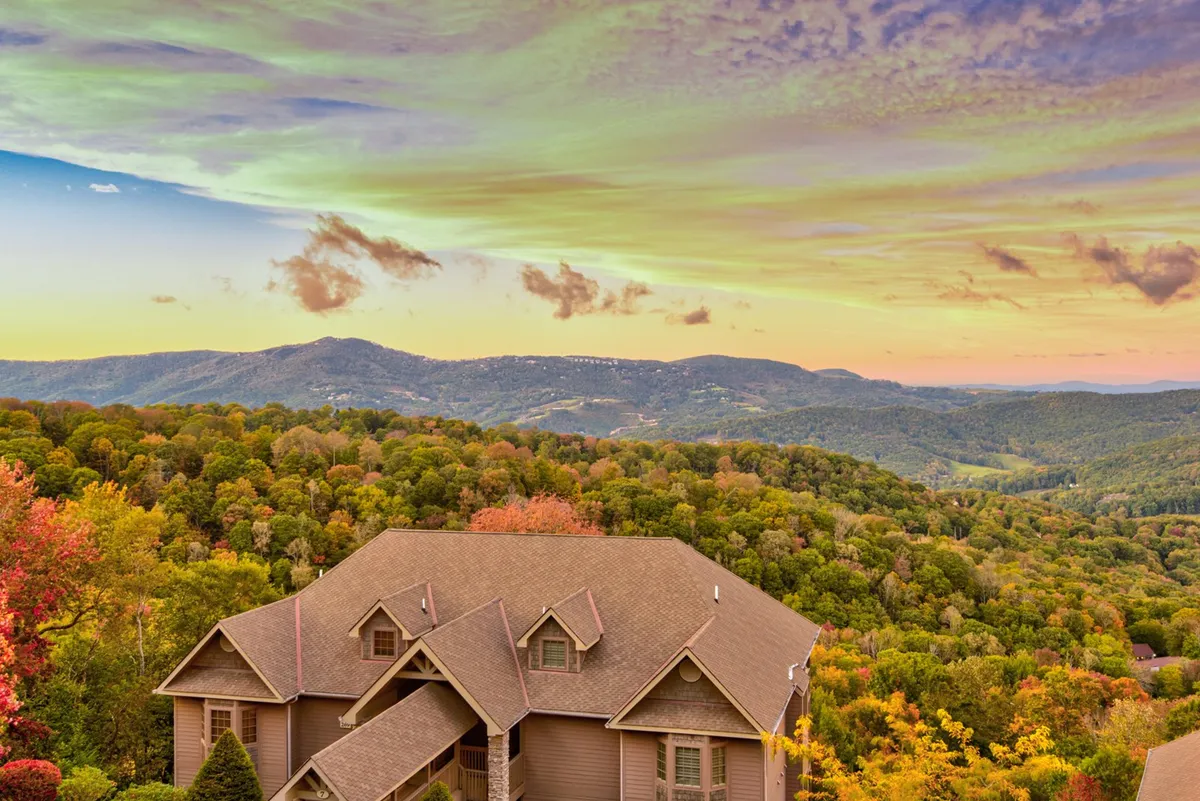 This exterior photograph features a luxurious house bordered by brilliant autumn trees and expansive mountain views, set beneath a soft, glowing sky. The inviting porch and surrounding landscape highlight opportunities for relaxation and exploration amid stunning natural beauty.